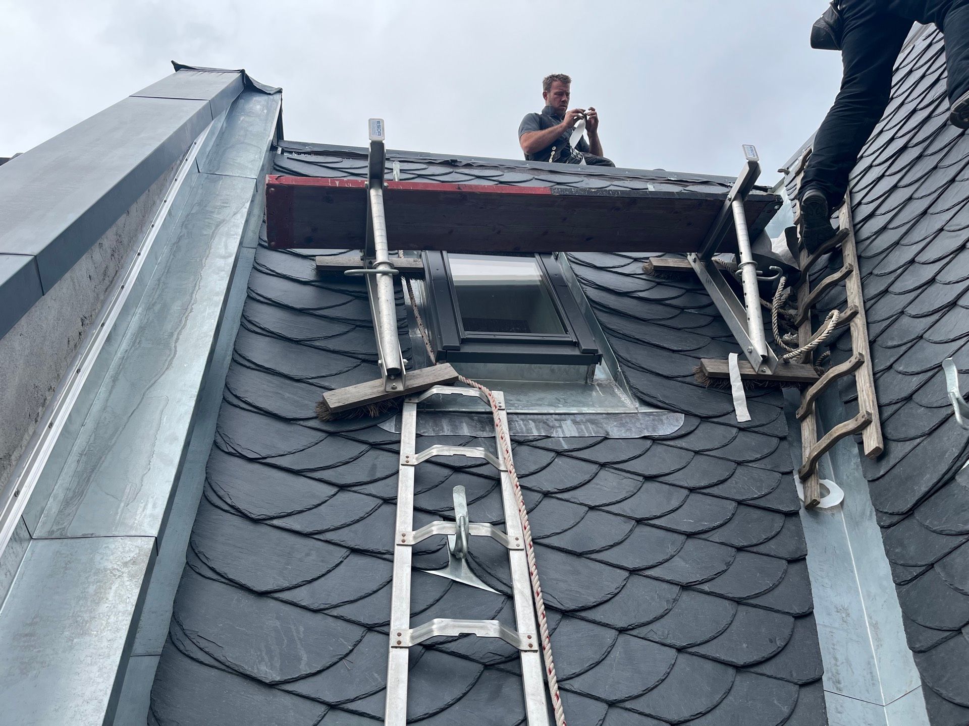 Dachdecker arbeitet mit Leiter und Sicherheitsausrüstung auf einem dunklen Schindeldach. Bewölkter Himmel.