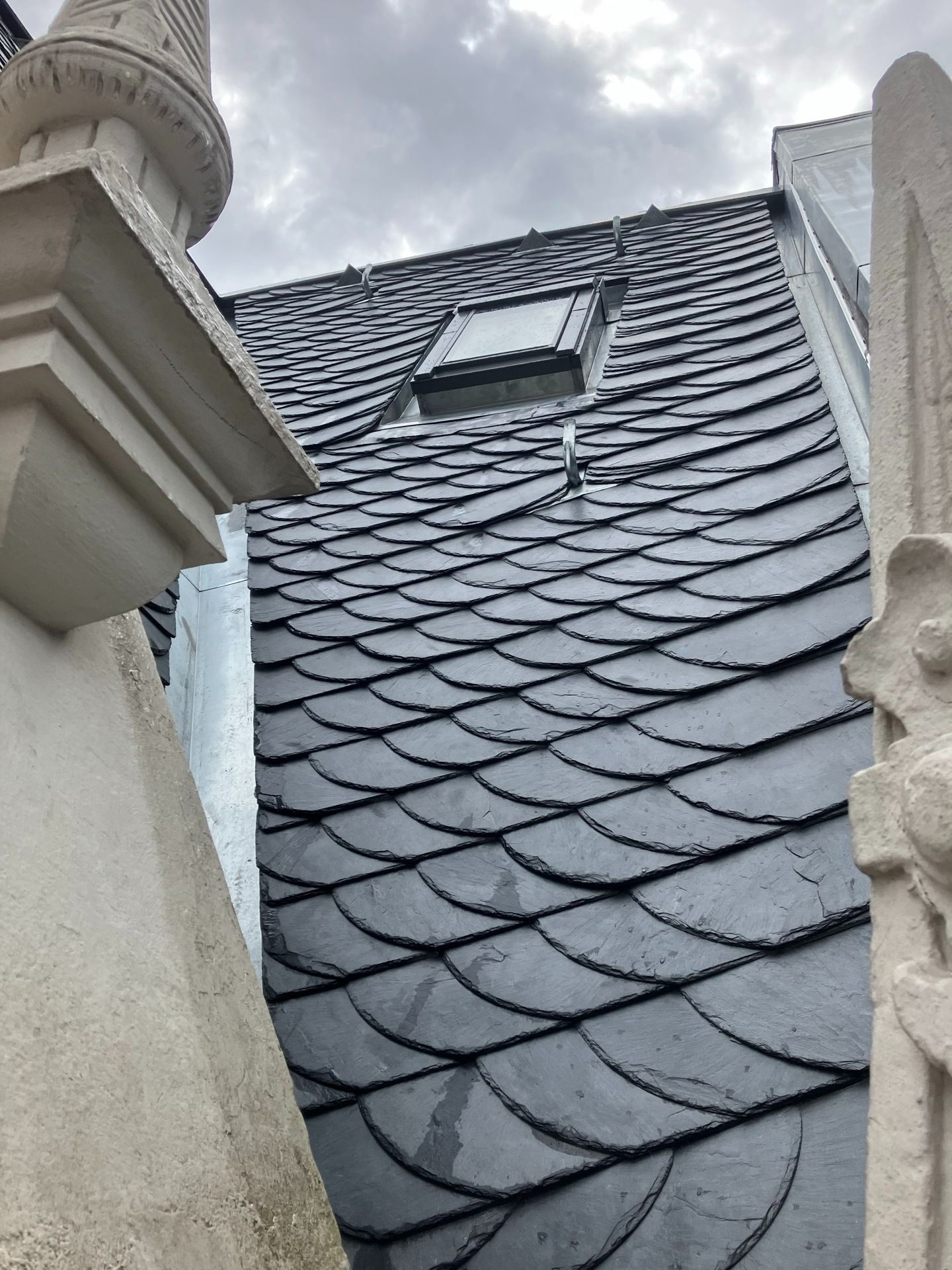 Dunkles Schieferdach mit Oberlicht unter bewölktem Himmel. Zwischen Steinsäulen zu sehen.