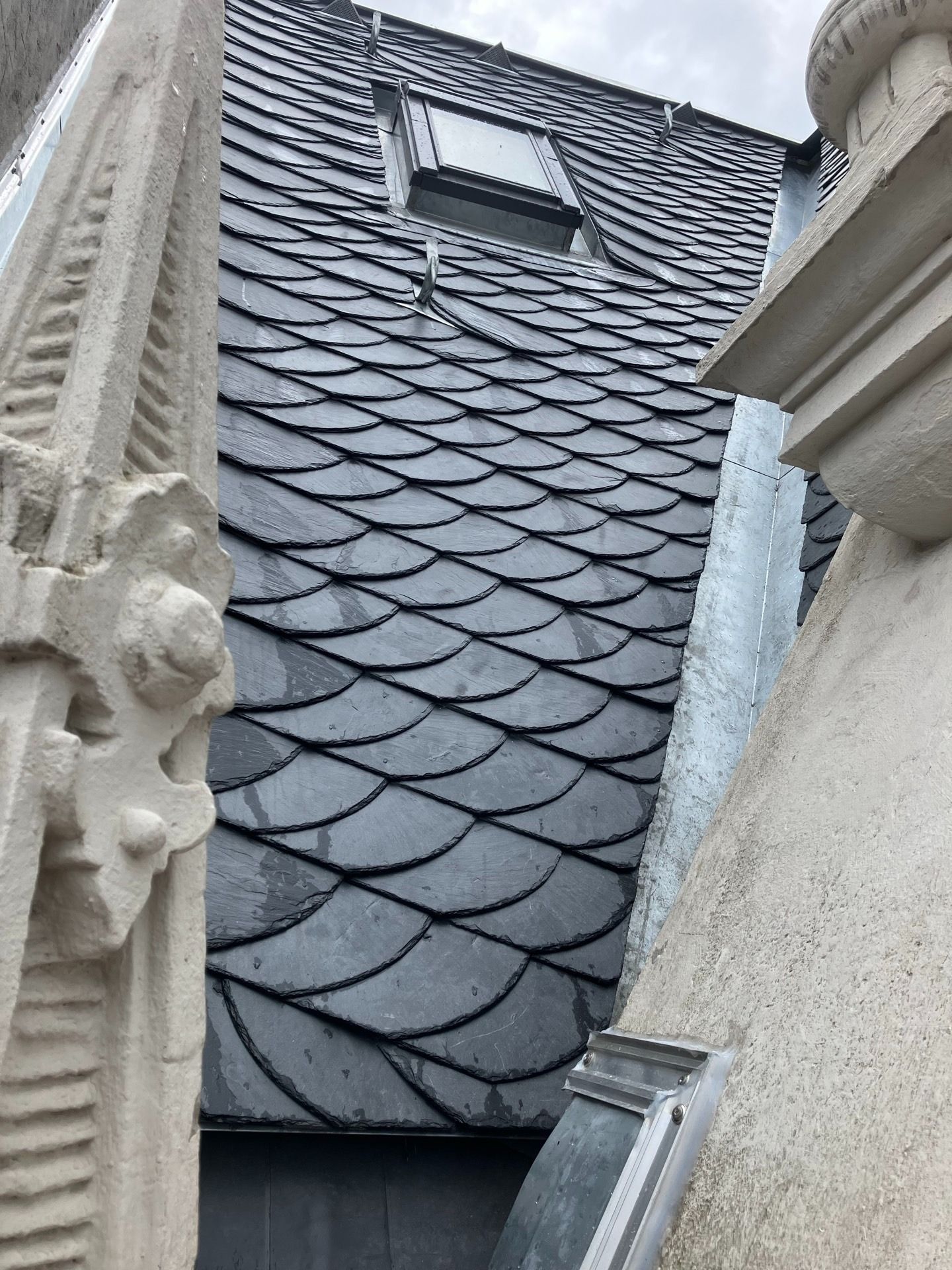 Schieferdach mit einem kleinen Fenster, das zwischen kunstvollen architektonischen Steinelementen zu sehen ist.