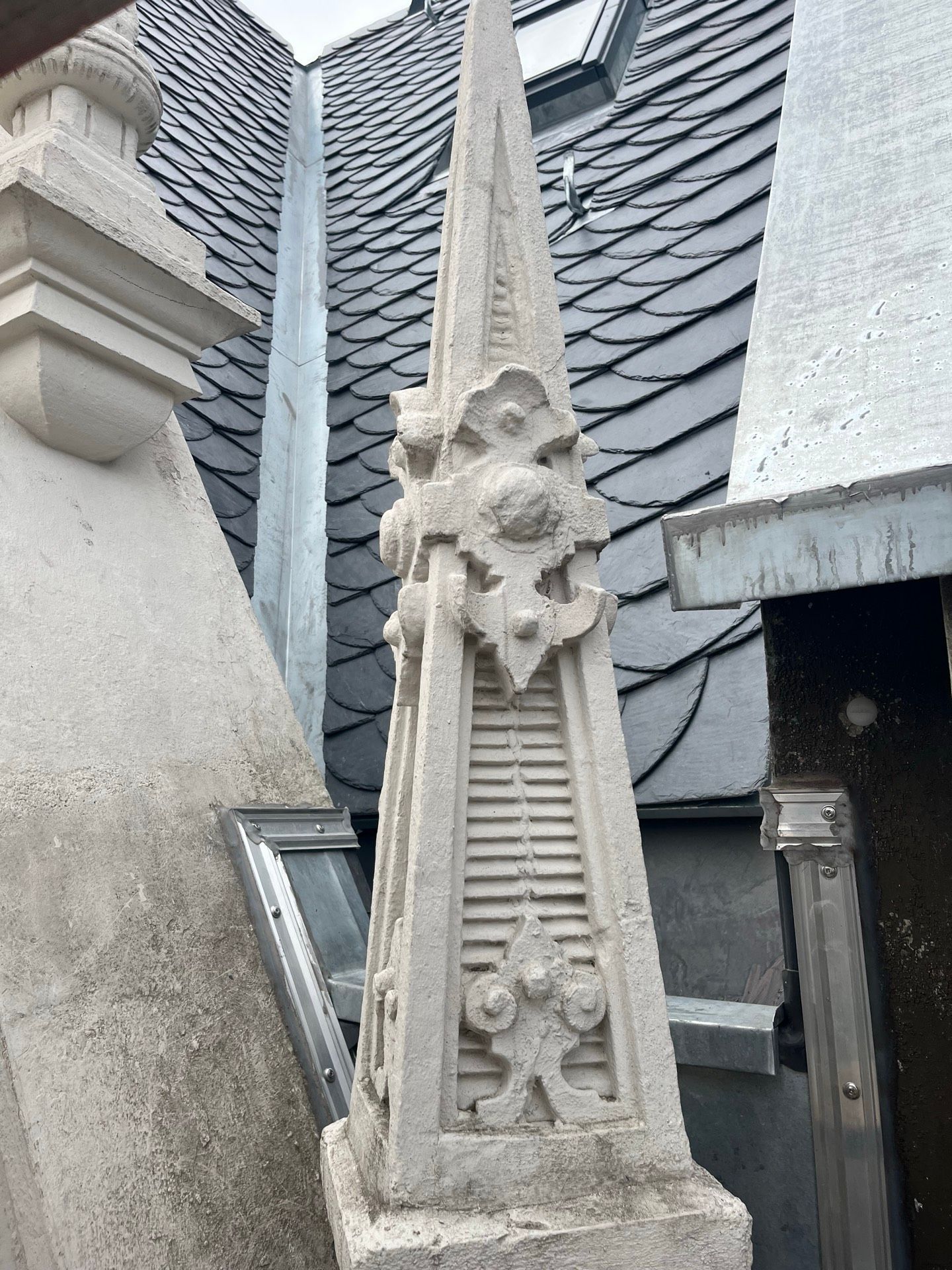 Verzierter, hellgrauer Turm mit dekorativen Elementen vor dunklen Dachziegeln.