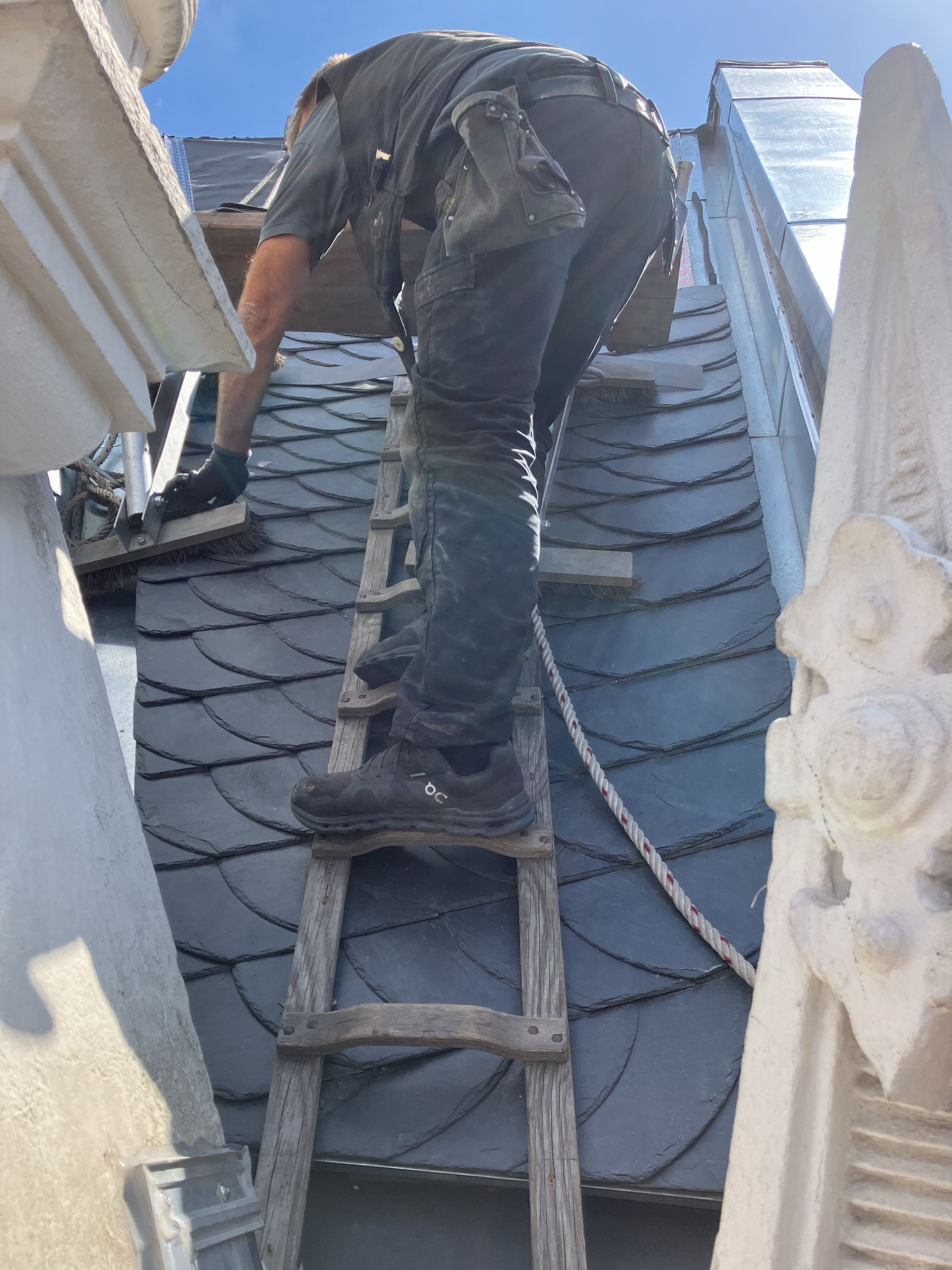 Person auf einer Leiter, die Schieferdachziegel verlegt. Graues Dach und Leiter. Strahlender Himmel.