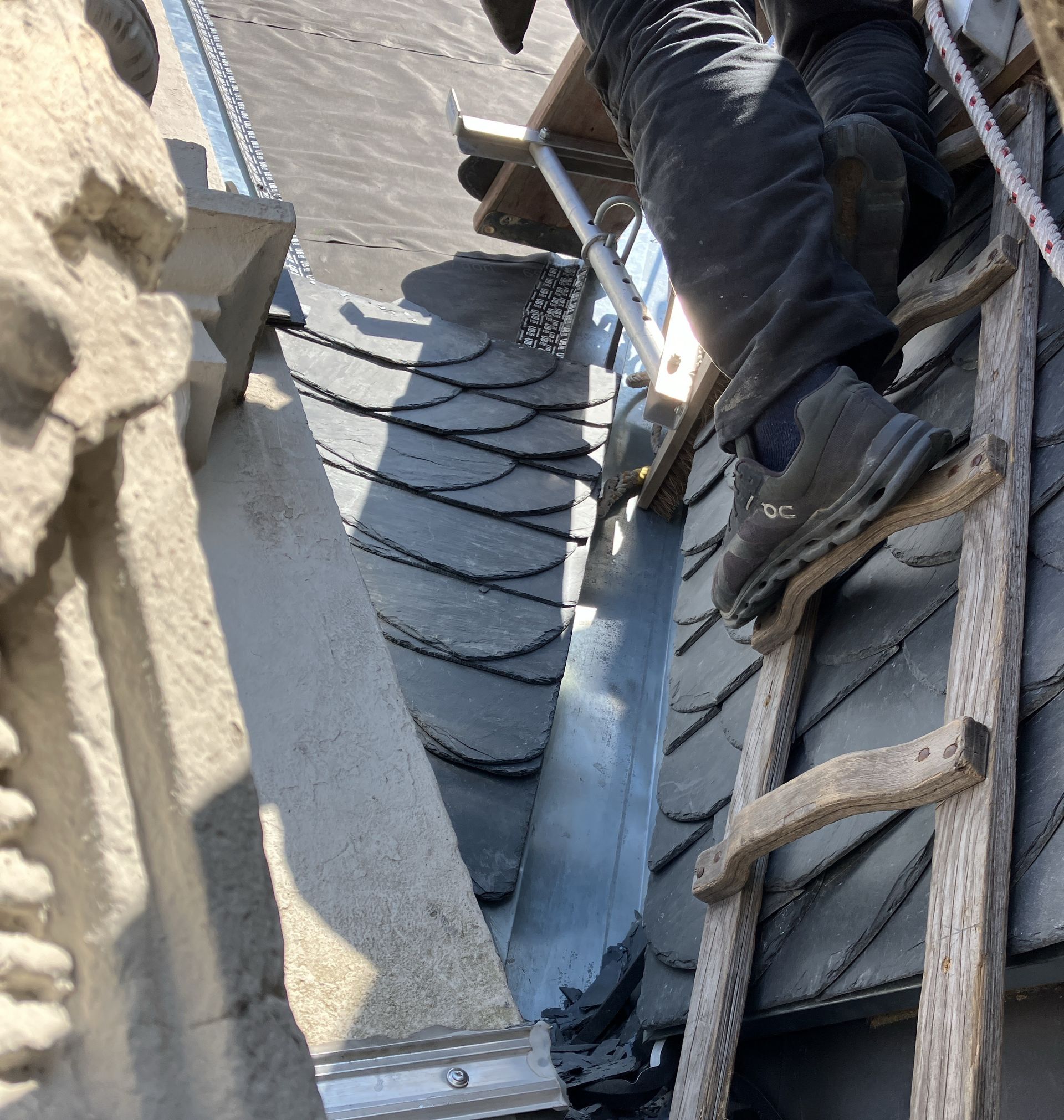 Person, die über eine Holzleiter auf ein Schieferdach neben der Steinfassade eines Gebäudes klettert.