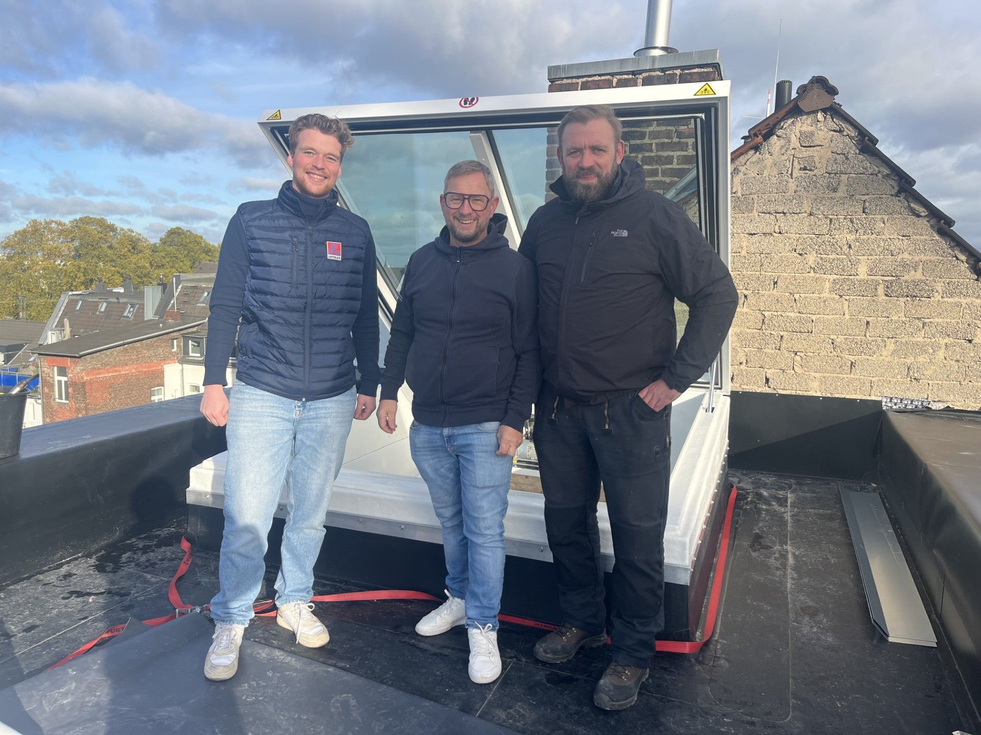 Three men stand on a flat roof next to a skylight. Cloudy sky in the background.