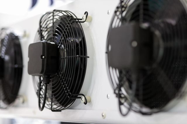 Fila de ventiladores negros montados en la pared en una habitación industrial luminosa.