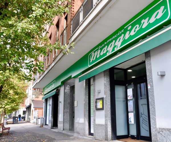 Farmacista in camice bianco sorride, braccia incrociate, in una farmacia con scaffali pieni di medicinali.