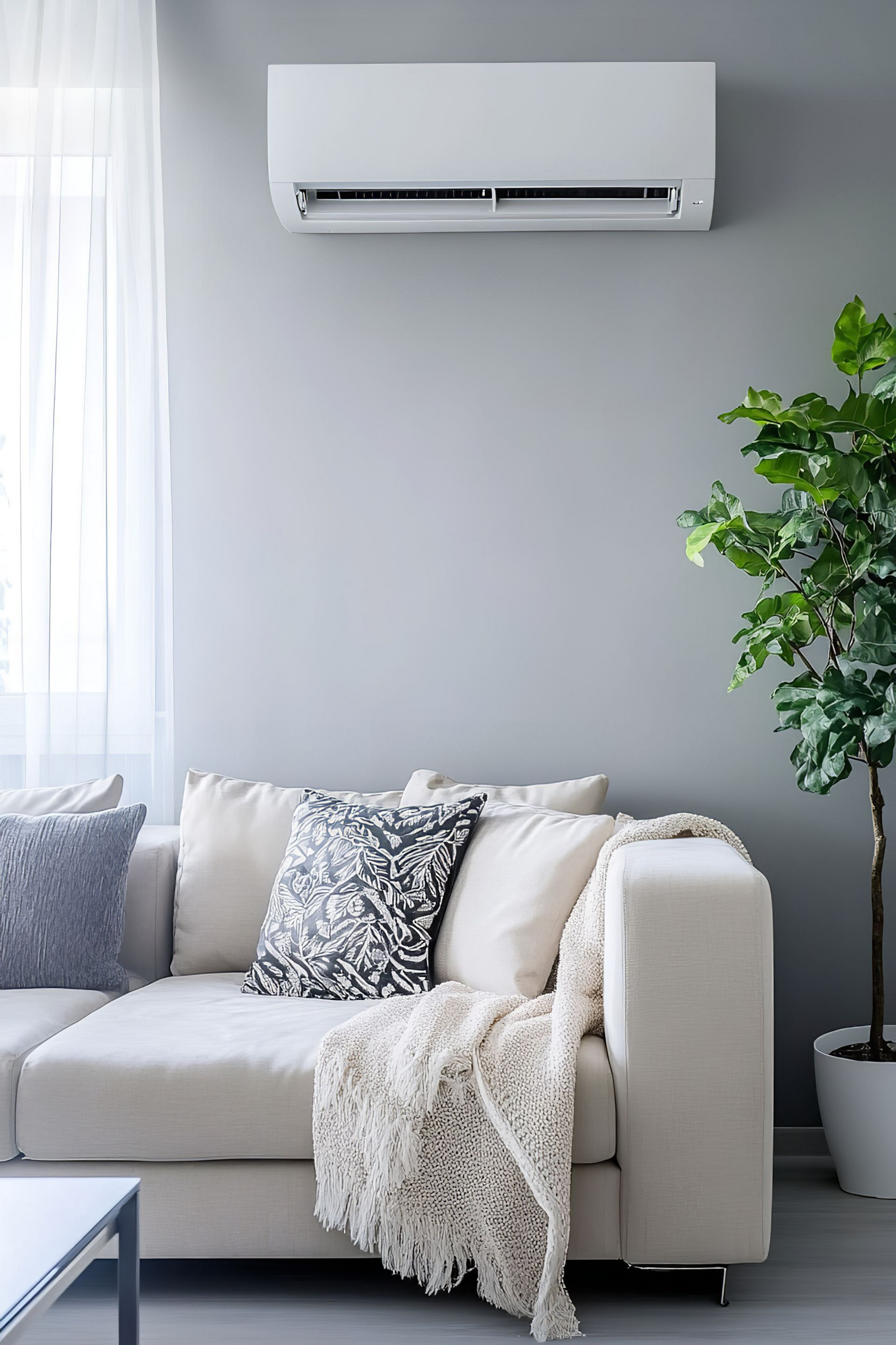 Un climatiseur est installé au-dessus d'un canapé beige agrémenté de coussins décoratifs, d'un plaid et d'une plante en pot dans un salon.