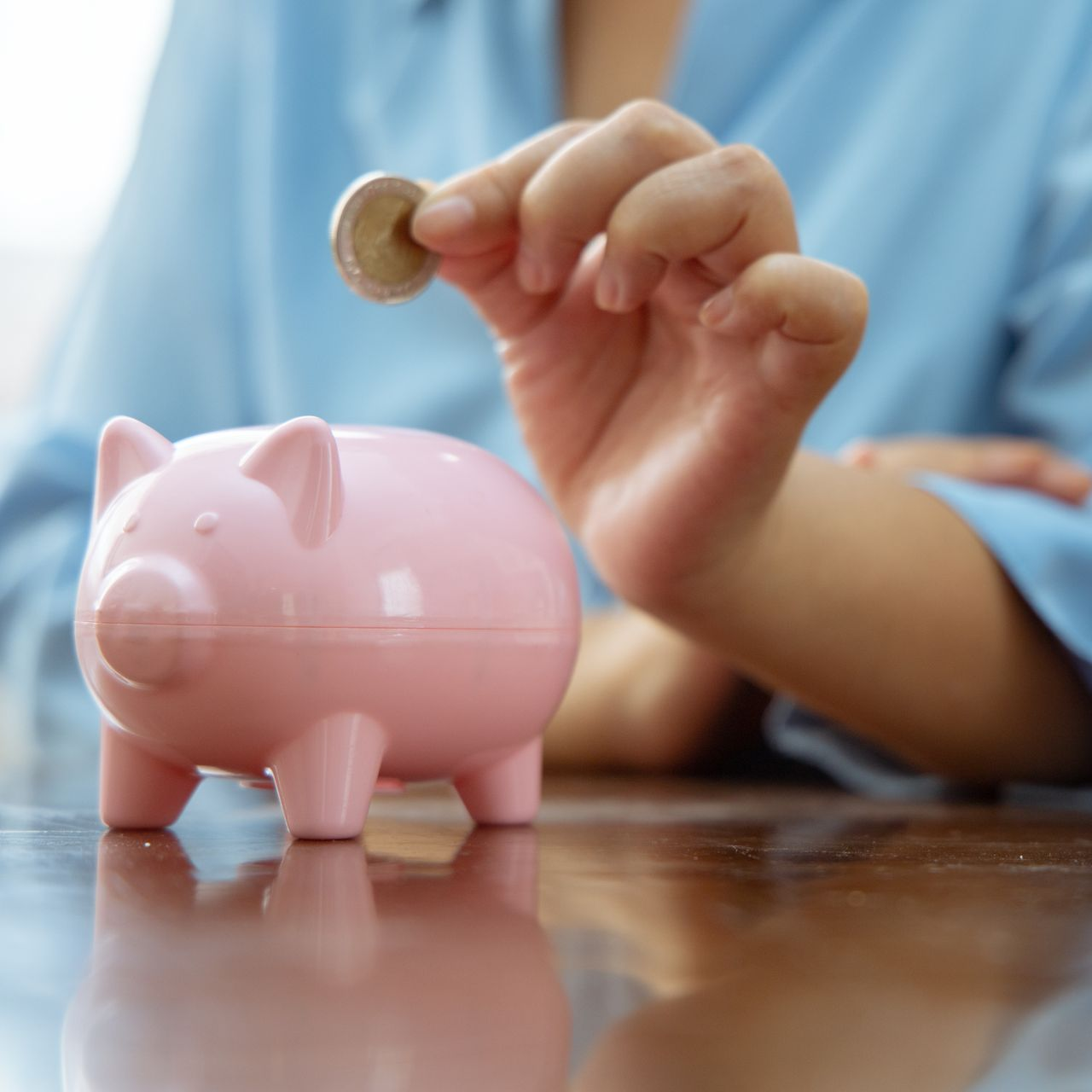 Une personne dépose une pièce dans une tirelire rose posée sur une table en bois.
