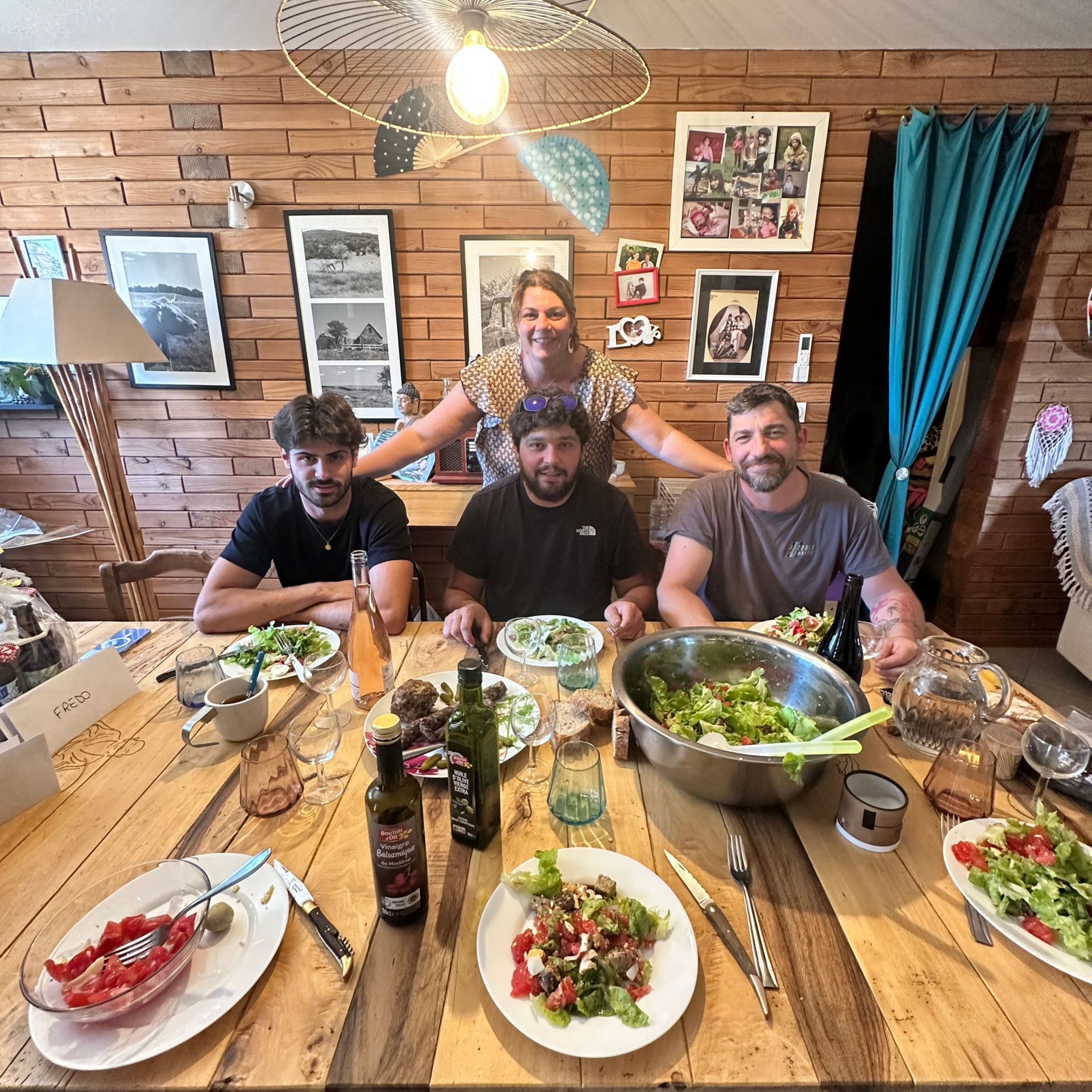 Cinq personnes attablées à une table en bois dégustent une salade. Une femme, souriante, se tient derrière elles. Intérieur rustique.