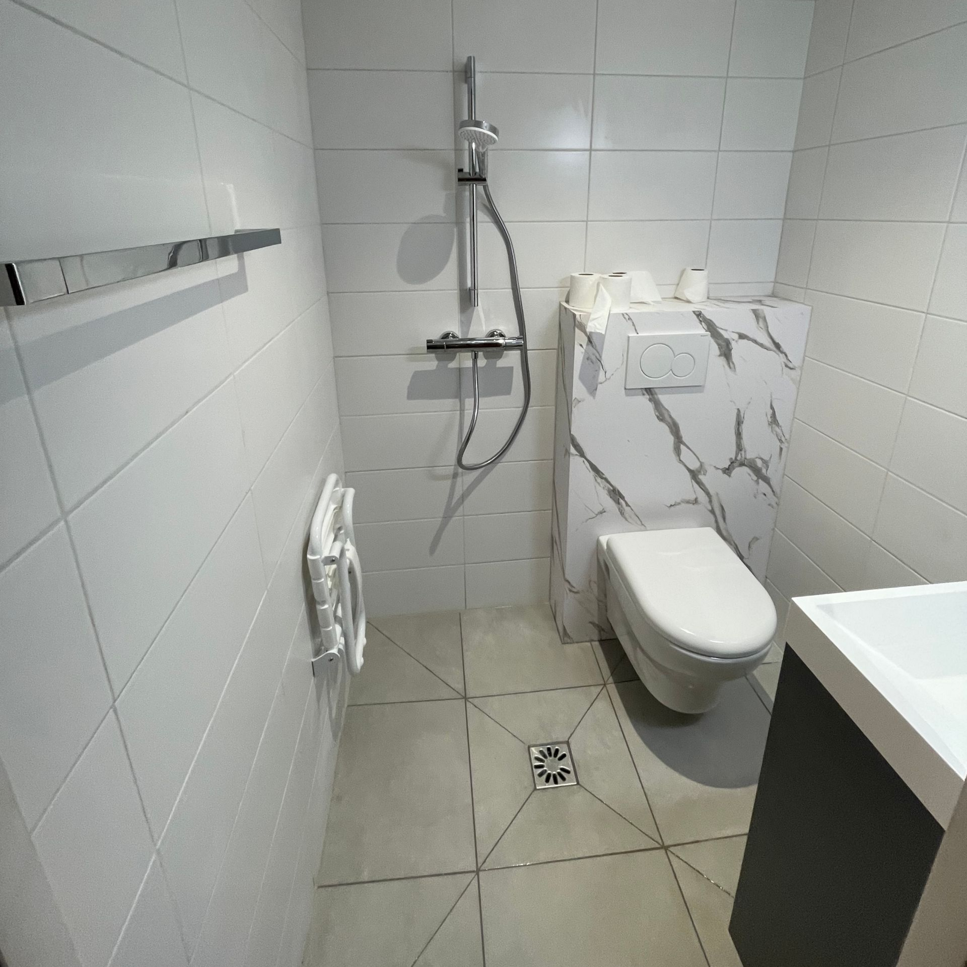 Petite salle de bains blanche avec toilettes suspendues, douche et lavabo.