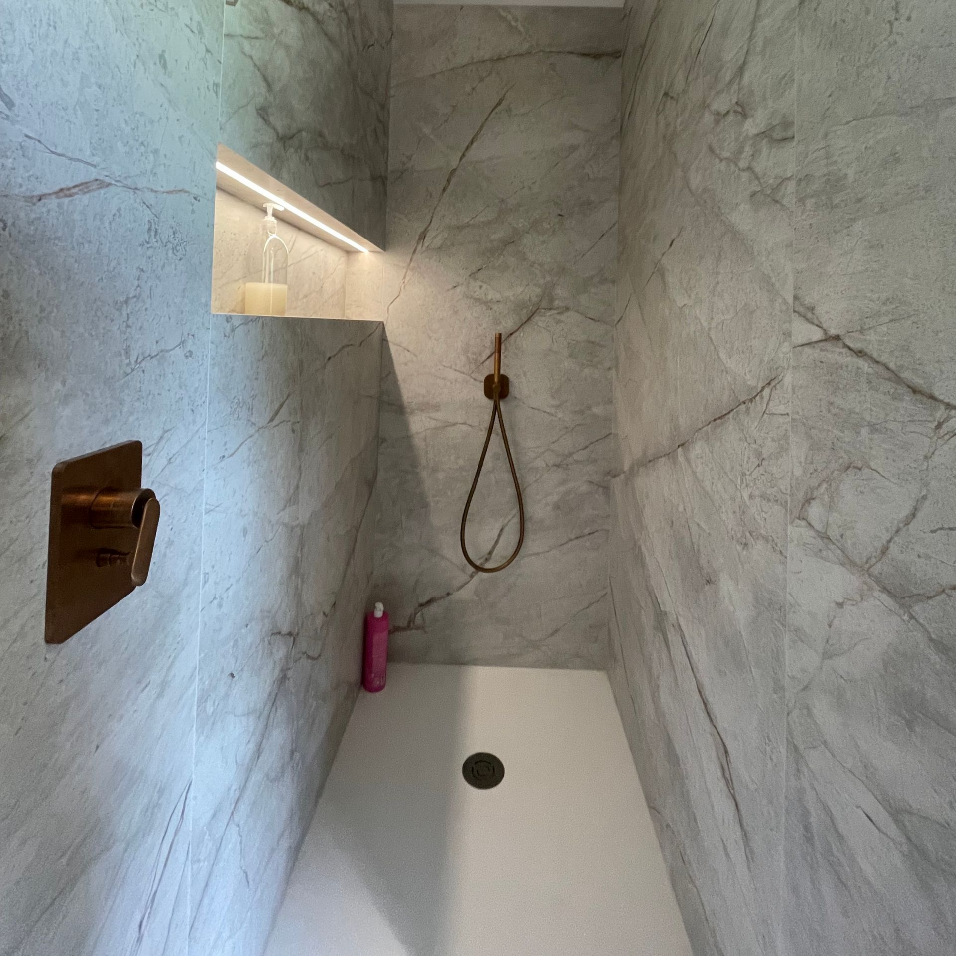 Douche avec carrelage gris imitation marbre, robinet doré, étagère intégrée avec éclairage et receveur blanc.