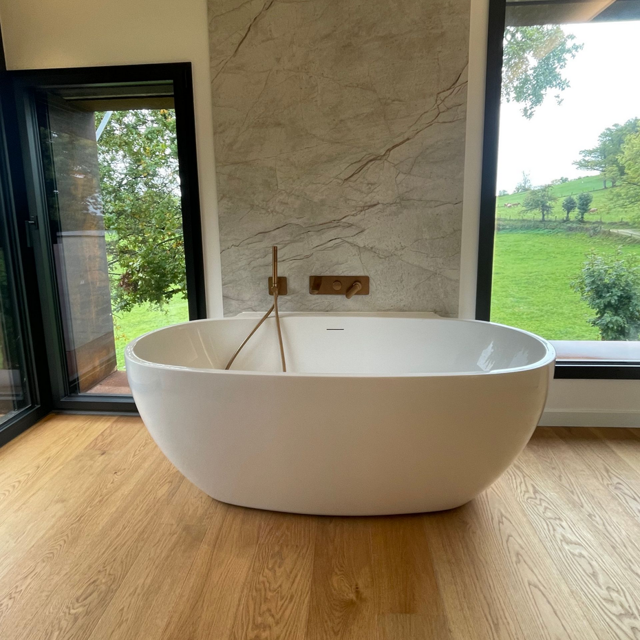 Baignoire ovale blanche près d'une fenêtre donnant sur une pelouse verdoyante. Robinetterie dorée et mur en marbre.