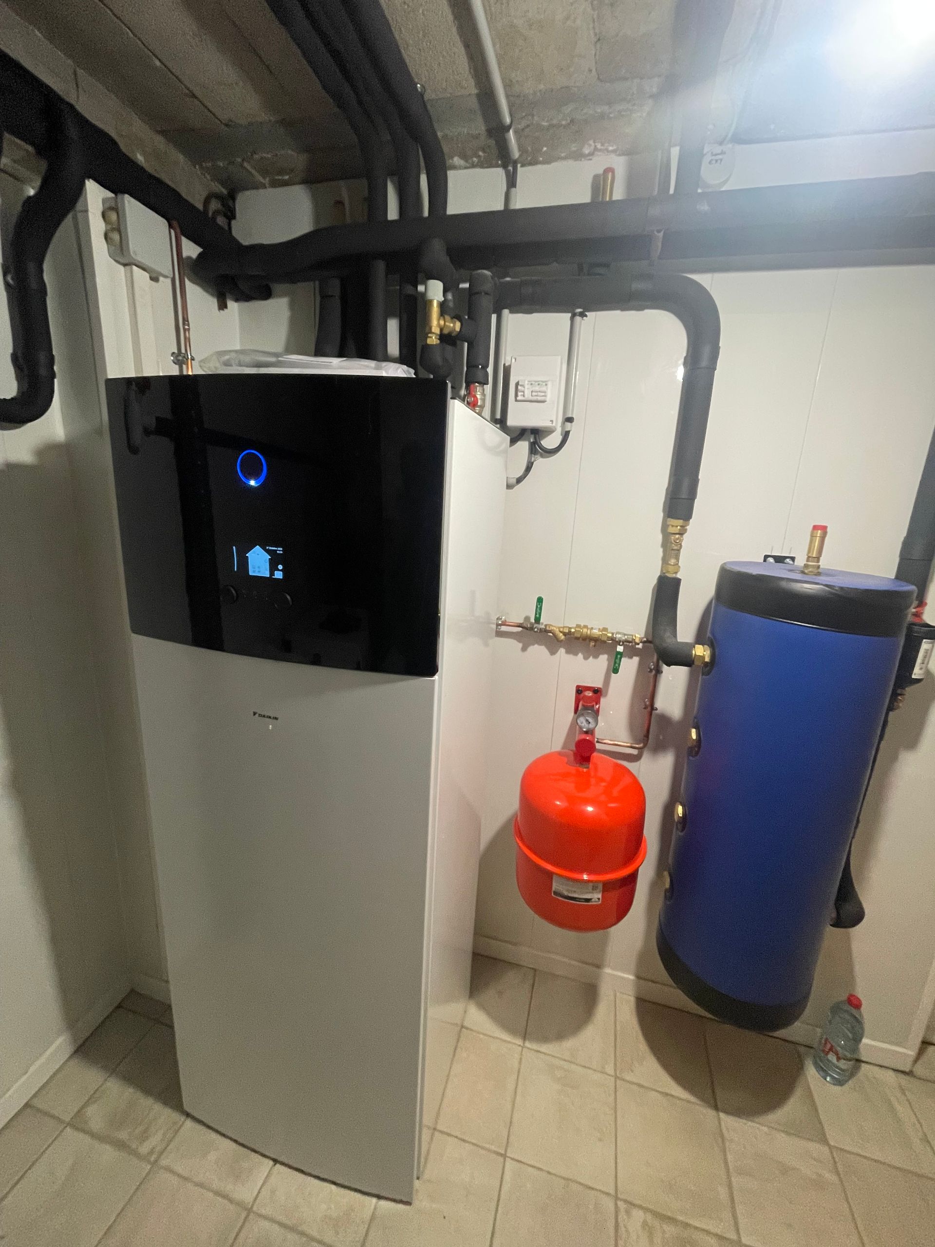 Système de pompe à chaleur avec une unité noir et blanc, un réservoir bleu et un vase d'expansion rouge dans une buanderie.