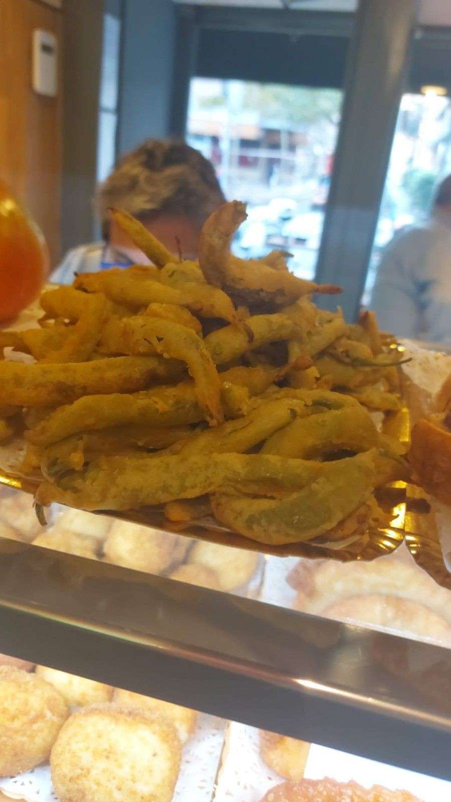 Pimientos verdes fritos apilados en una bandeja en una vitrina de delicatessen, con una persona visible detrás de ellos.