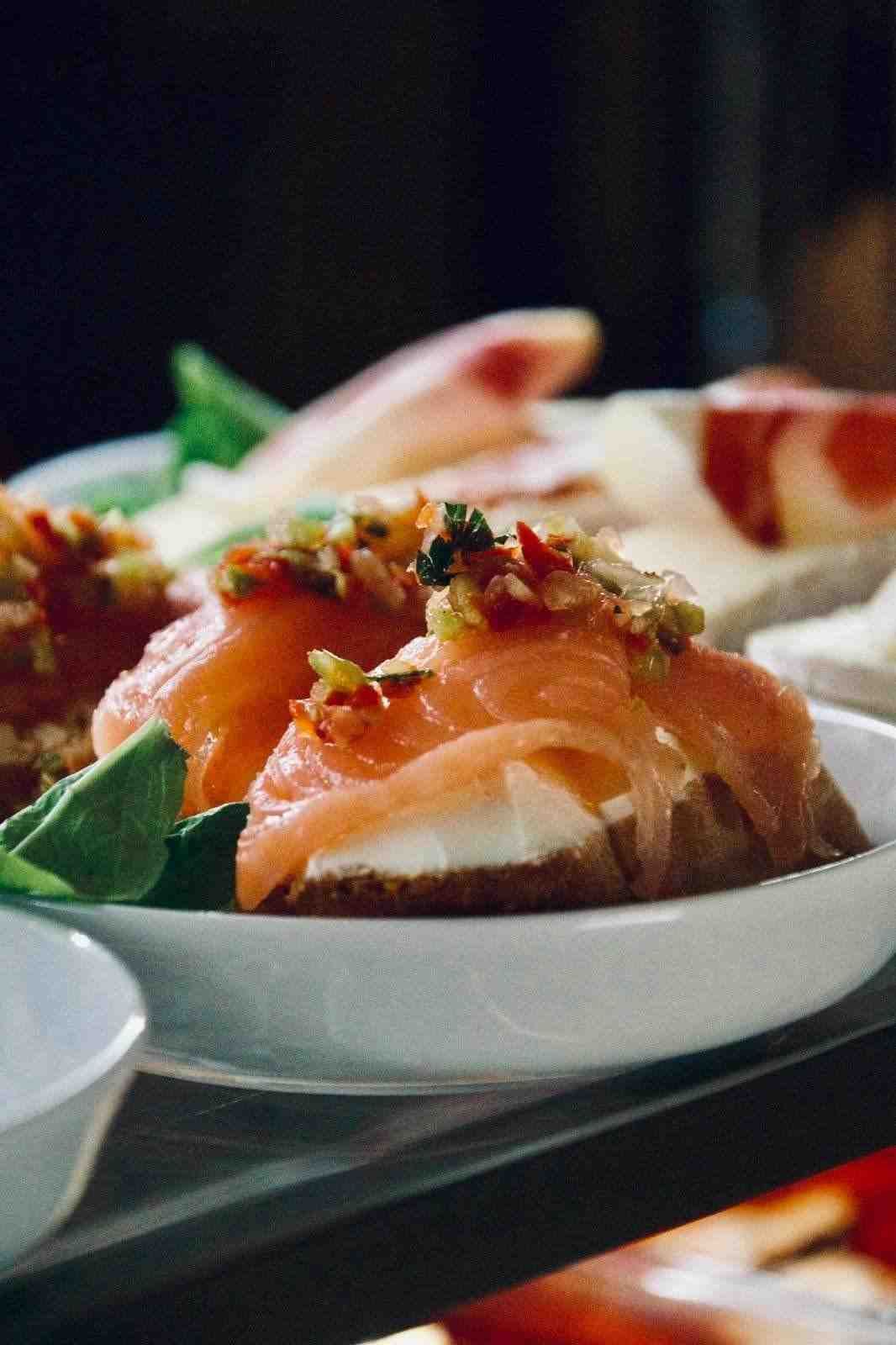 Aperitivos de salmón ahumado en un plato blanco, cubiertos con salsa, guarnición de albahaca y otros aperitivos de fondo.