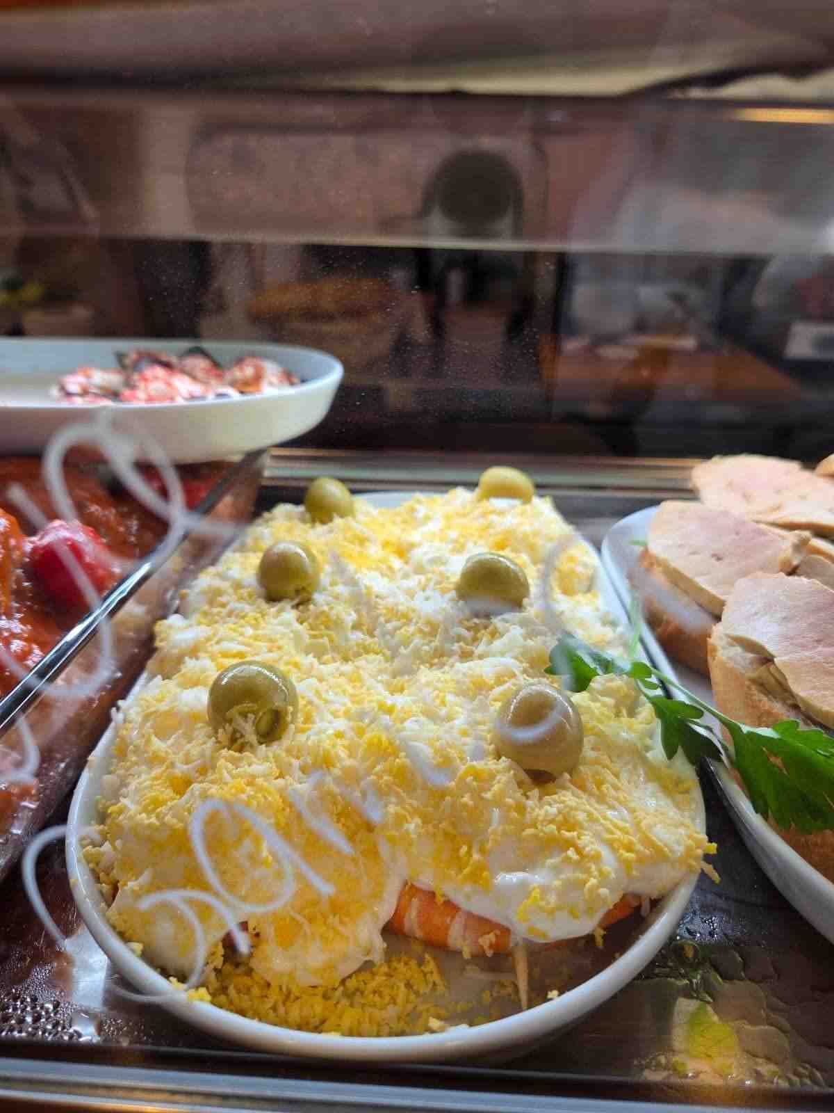 Plato ovalado de ensalada de patatas con aceitunas y huevo rallado, con otras tapas visibles.