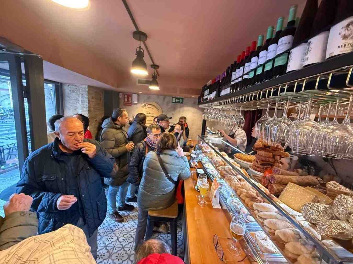 Gente en un bar con aperitivos, bebidas y botellas de vino. Una persona está comiendo.