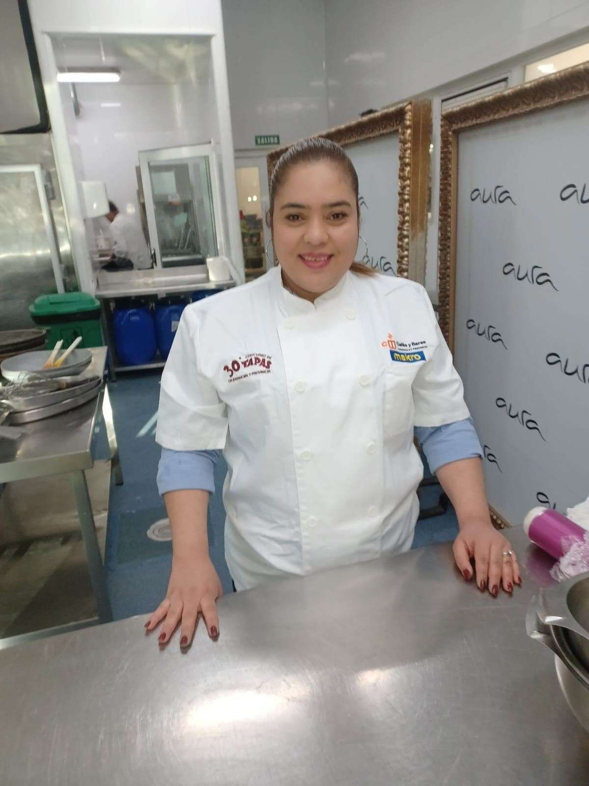 Una mujer con chaqueta de chef sonríe en una cocina de acero inoxidable.
