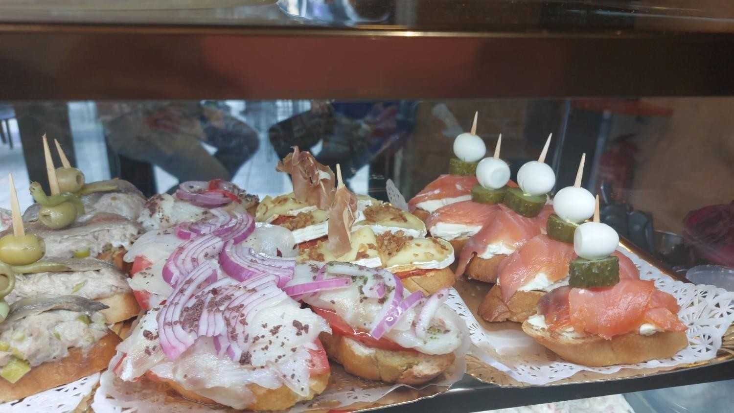 Exposición de diversas tapas sobre rebanadas de pan, incluyendo pescado, cebolla y queso, en una vitrina de cristal.