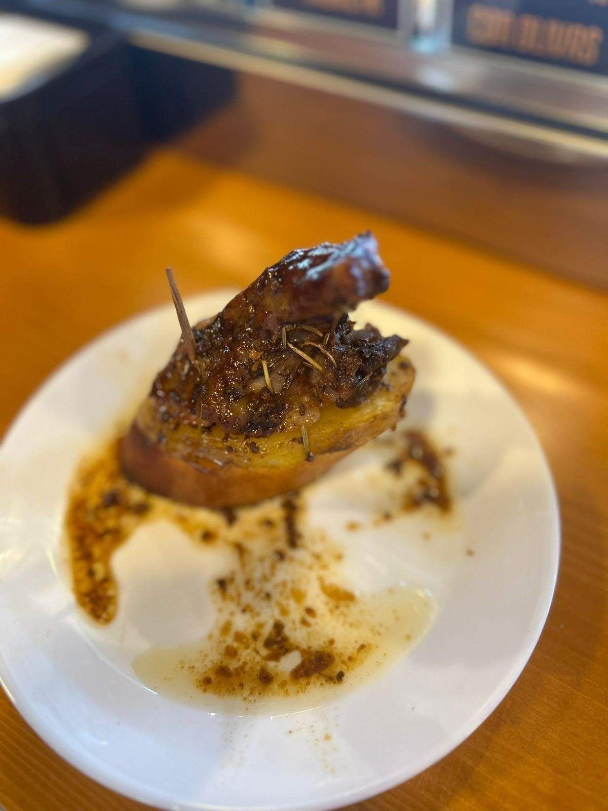 Una papa cocida cubierta con carne oscura y salsa en un plato blanco, mesa de madera.