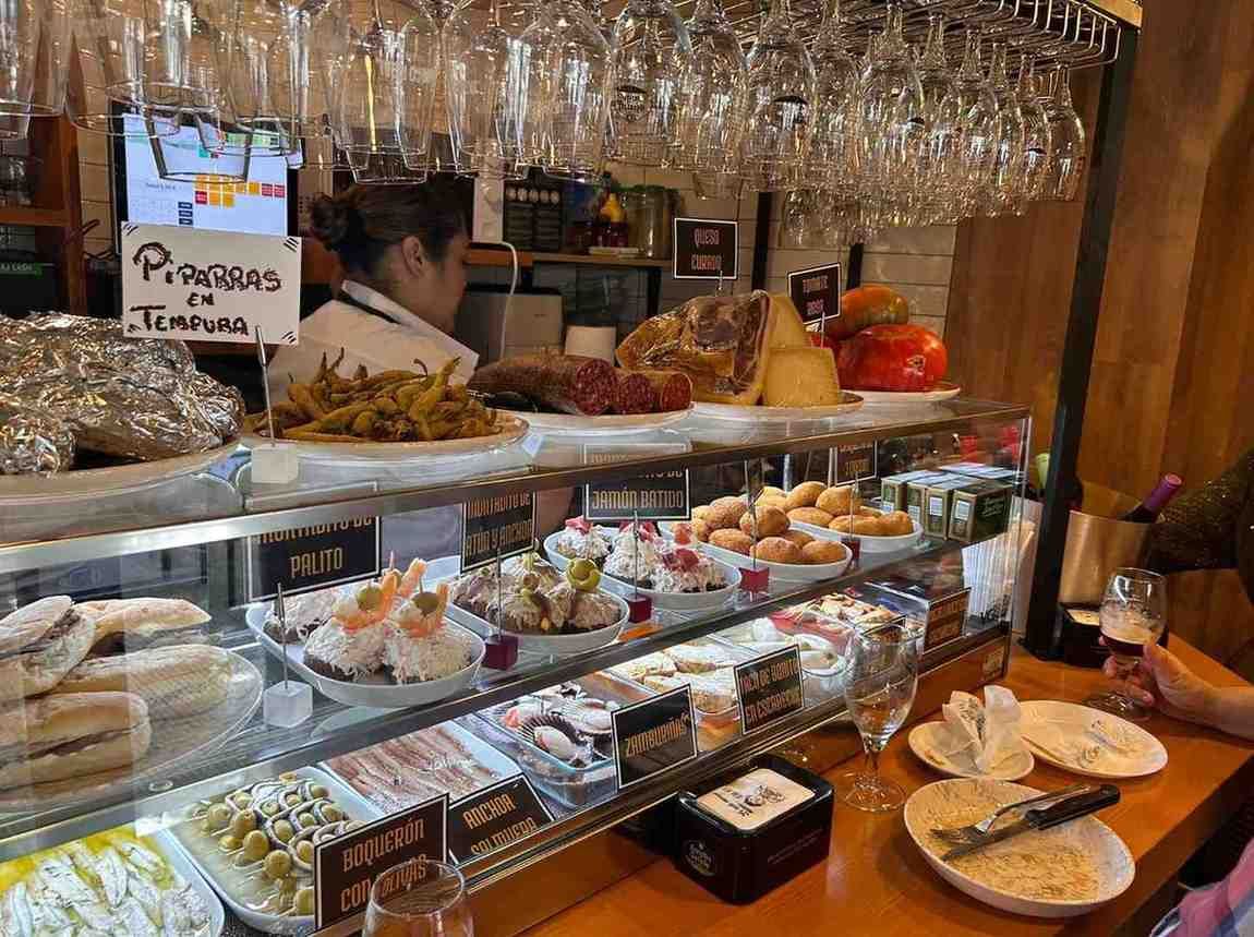 Una vitrina de comida en un bar, llena de tapas variadas. Un camarero está detrás del mostrador.