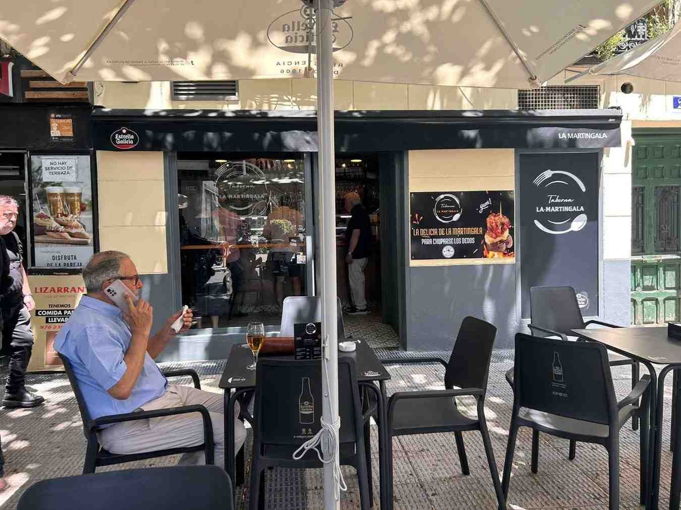 Una persona hablando por teléfono en una mesa fuera de un bar, con otras personas visibles dentro. 
