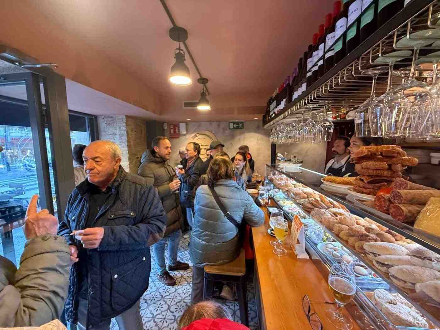 Interior de bar con clientes; mostrador de madera, exhibición de tapas, personal de bar, botellas de vino y luces colgantes.
