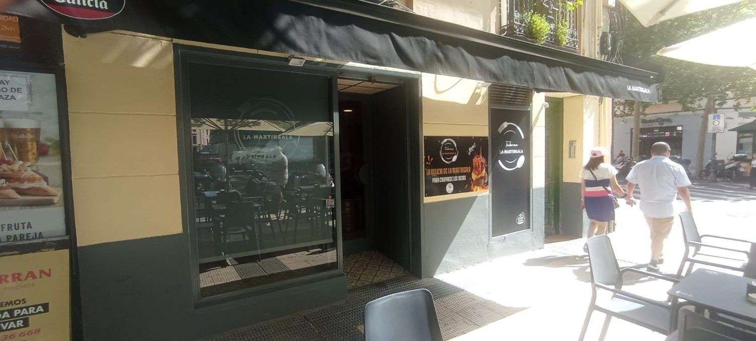 Exterior de un restaurante con toldo negro y la puerta abierta. Dos personas pasan. Mesas al aire libre.