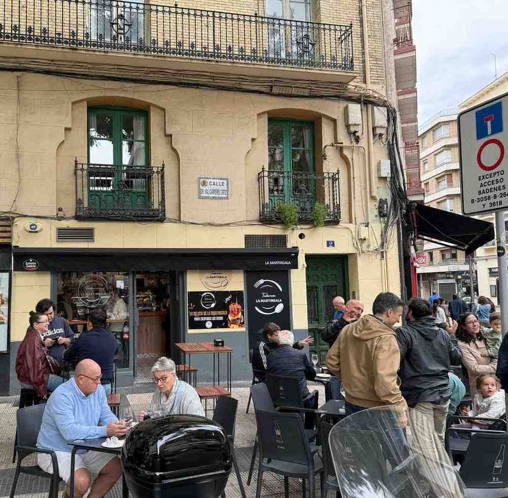 Gente en una cafetería con terraza. El edificio es amarillo con puertas y balcones verdes.