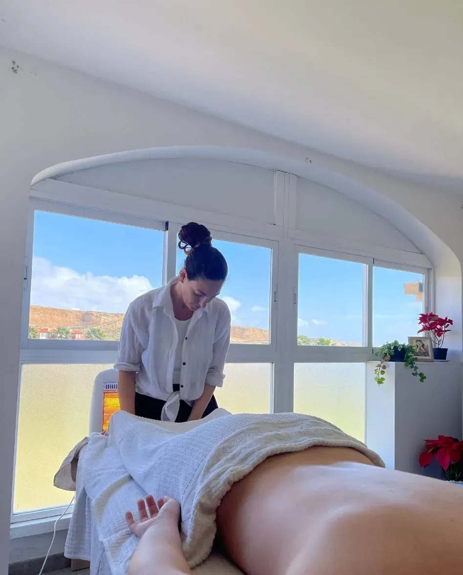 Mujer dando un masaje en el interior, sobre una persona acostada en una mesa de masajes cerca de una ventana.