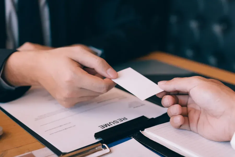 Dos manos intercambian tarjetas de visita sobre un escritorio con documentos y un cuaderno.