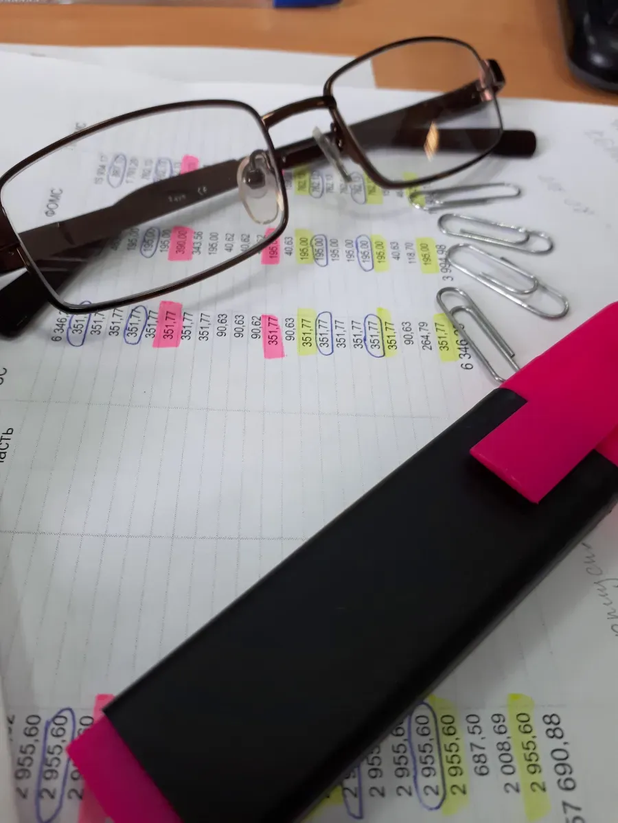 Gafas sobre un cuaderno con notas resaltadas, un bolígrafo y clips de papelería rosas y negros.