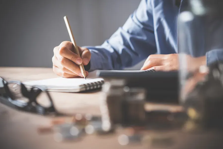 Dos personas revisan documentos al otro lado de un escritorio; una de ellas señala con un bolígrafo cerca de una computadora portátil y una calculadora.