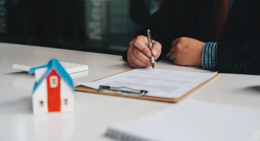 Firma a mano de un documento junto a una pequeña maqueta de una casa sobre un escritorio.