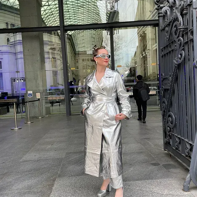 Una mujer con una gabardina plateada y gafas de sol está parada frente a un edificio.
