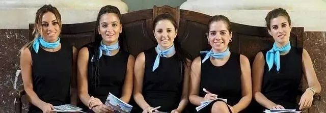 Un grupo de mujeres vestidas con vestidos negros y bufandas azules están sentadas una al lado de la otra.