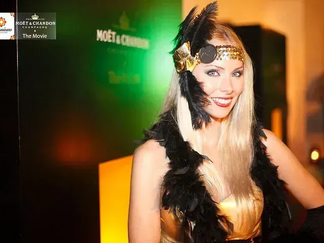 Una mujer que lleva una diadema y una boa de plumas está parada frente a un cartel de Moët & Chandon.