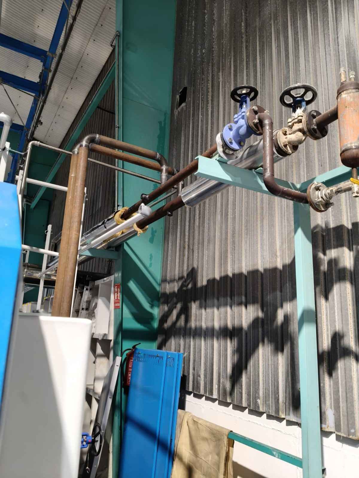 Tuberías y válvulas industriales junto a una pared de metal corrugado, con tuberías azules y de cobre en la parte superior.
