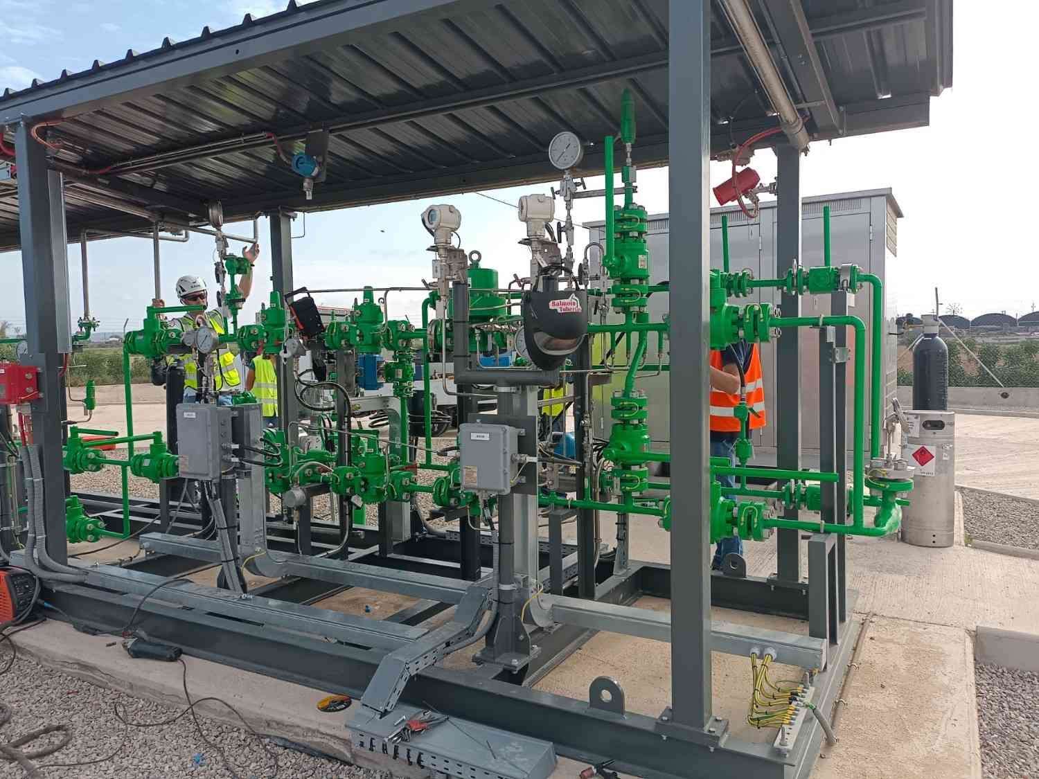 Tuberías y equipos industriales bajo una marquesina metálica, con válvulas y bombas verdes y grises en el exterior.