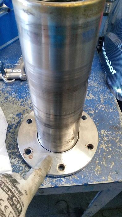 Cilindro de metal montado sobre una base circular blanca en un banco de trabajo azul, con una mano enguantada señalándolo.
