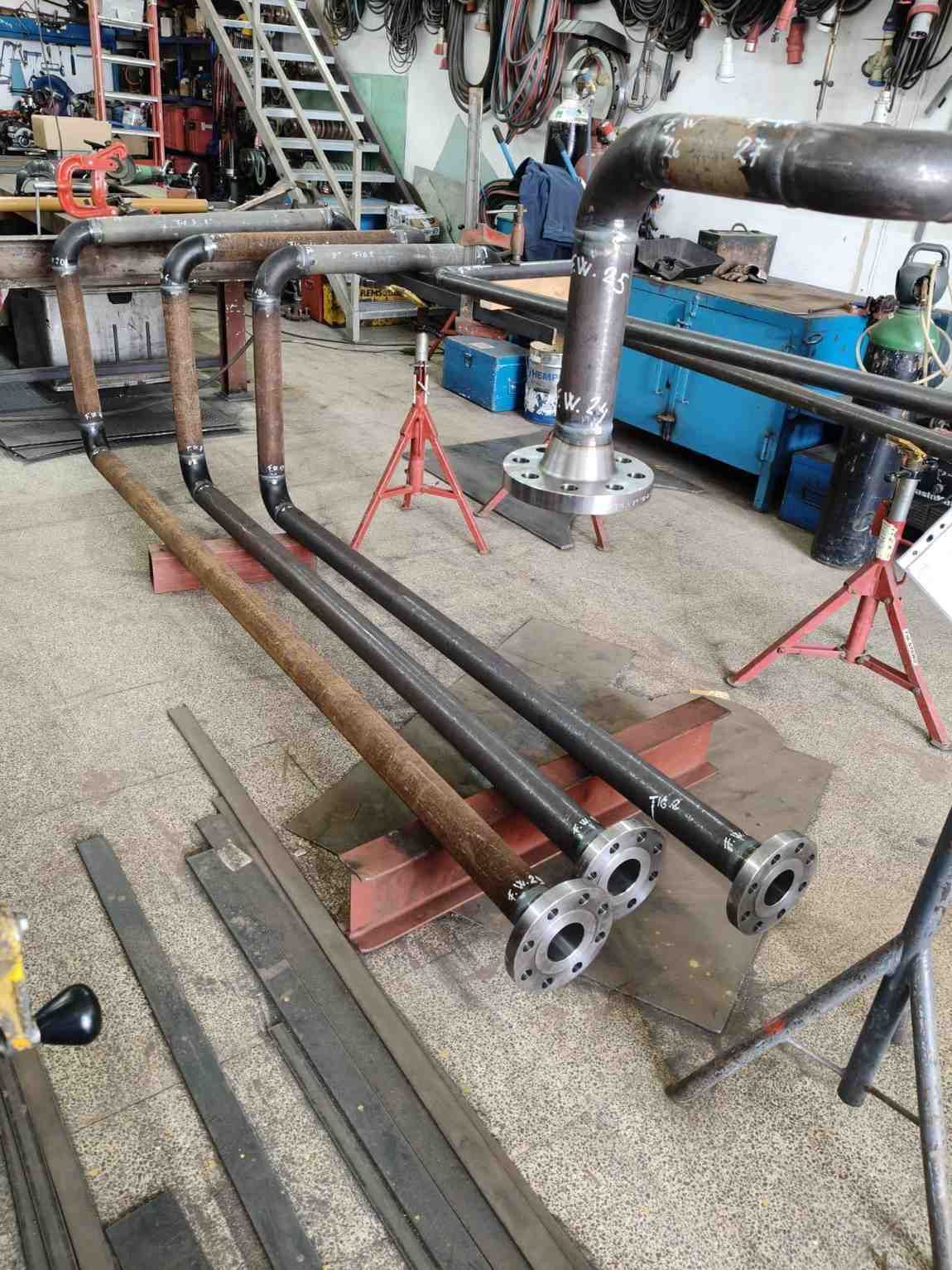 Grandes tuberías y bridas metálicas colocadas en el suelo de un taller industrial, con soportes rojos.