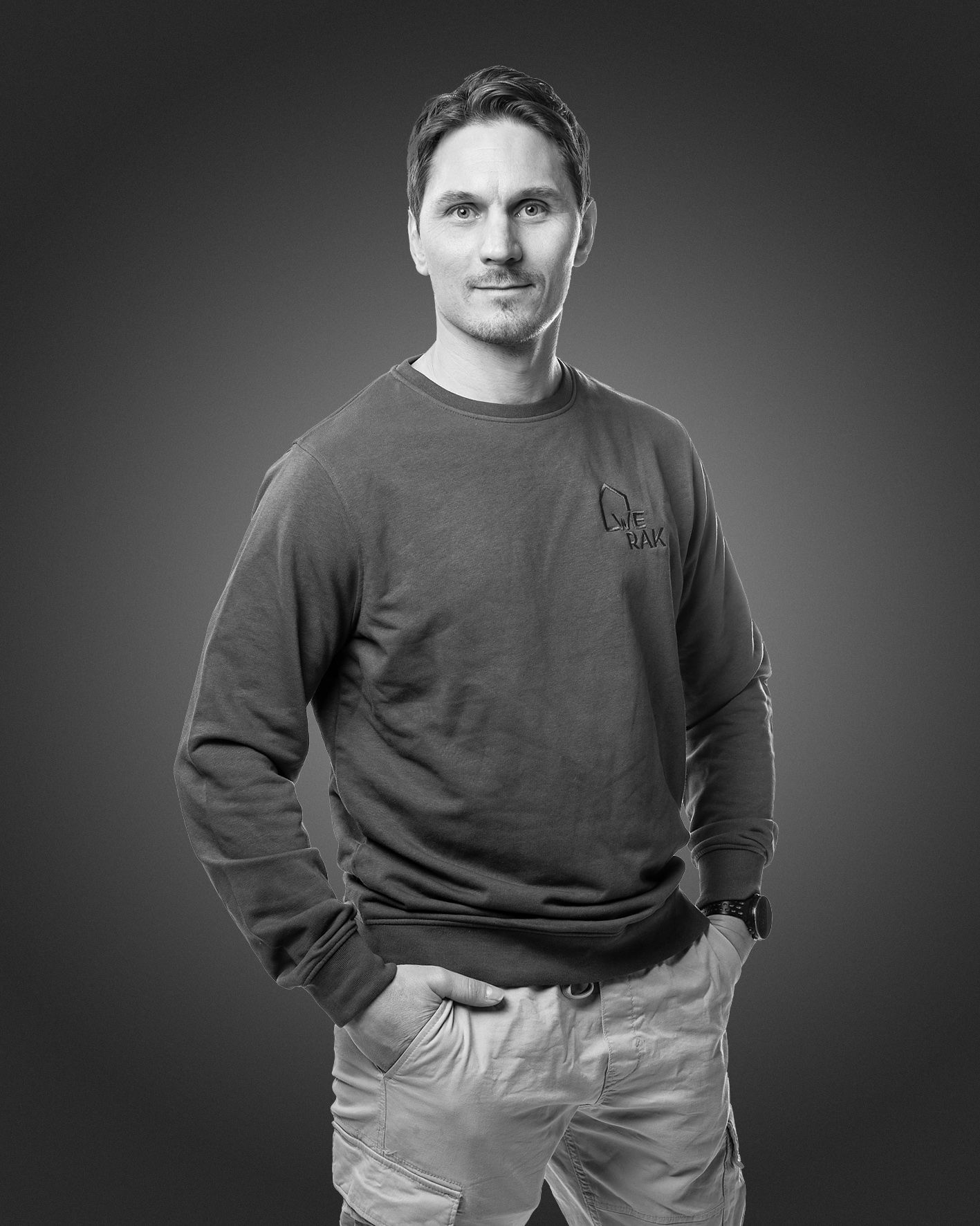 Tummaan neuleeseen ja vaaleisiin housuihin pukeutunut henkilö seisoo kädet taskuissa tummaa taustaa vasten.