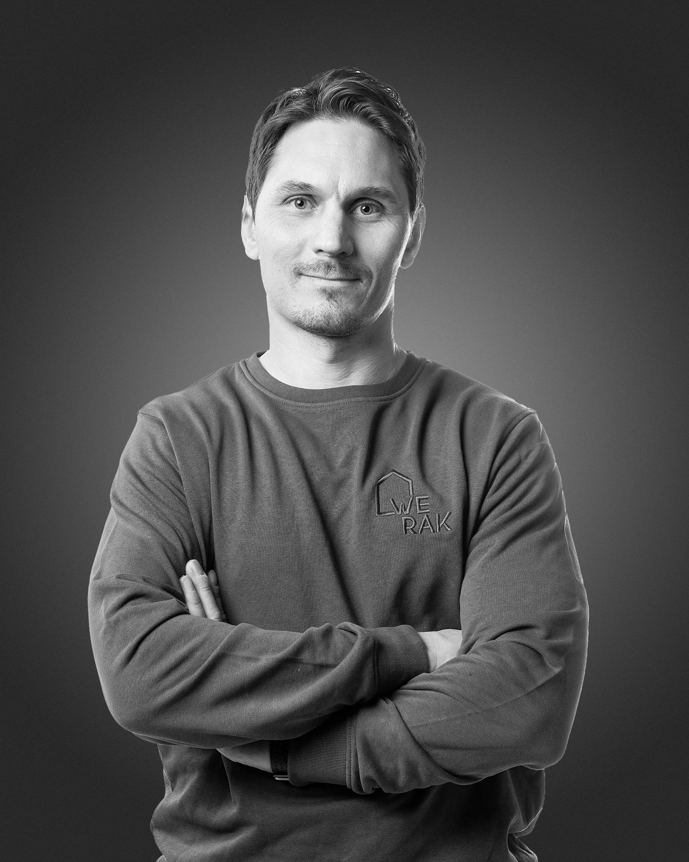 Lyhyet tummat hiukset ja vaalea parta, pitkähihainen paita, kädet ristissä, tummaa taustaa vasten.