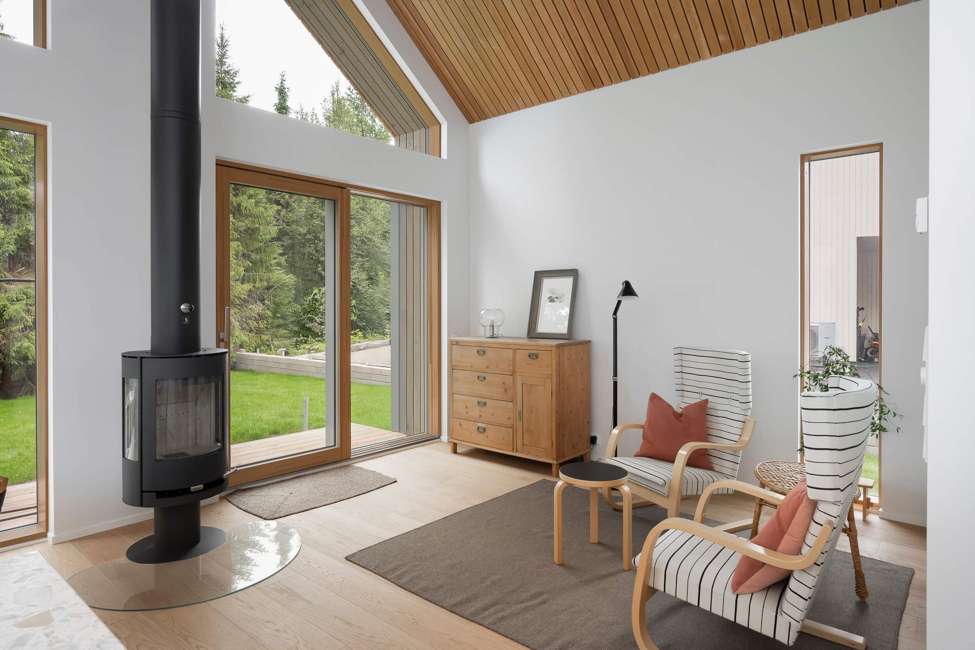 Kirkas ja moderni olohuone, jossa on puuhella, vaaleat puulattiat, puinen lipasto ja kaksi kuviollista nojatuolia.