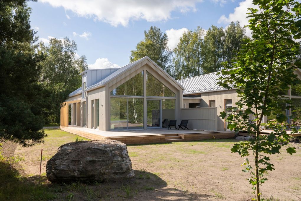 Moderni, vaaleanharmaa puutalo, jossa on suuri lasinen päätyseinä, aurinkoisessa, puiden reunustamassa maisemassa suuren kallion keskellä.