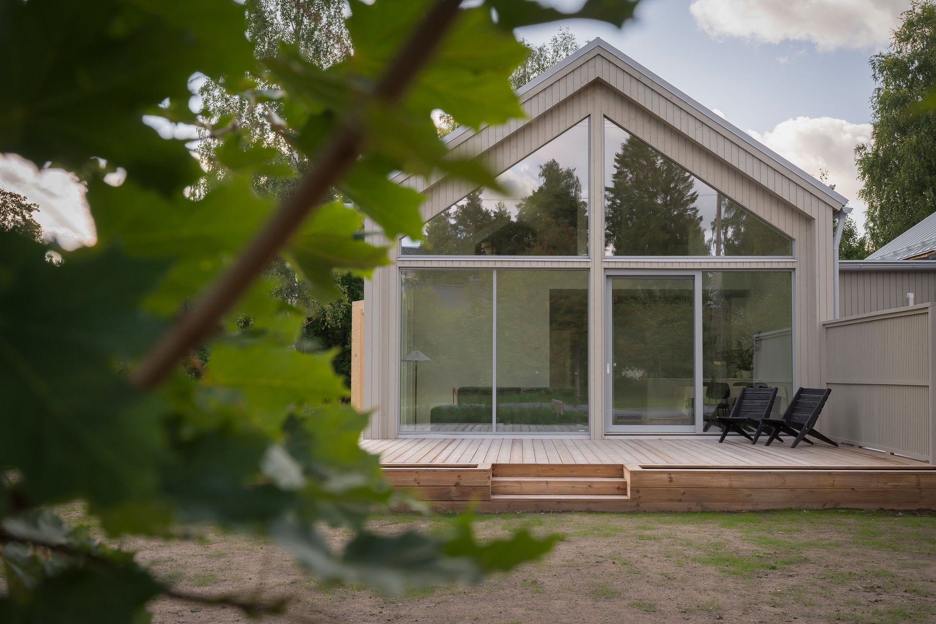 Moderni talo, jossa on harjakatto ja suuret lasi-ikkunat, puinen terassi ja kaksi lepotuolia nurmikkoisella pihalla.