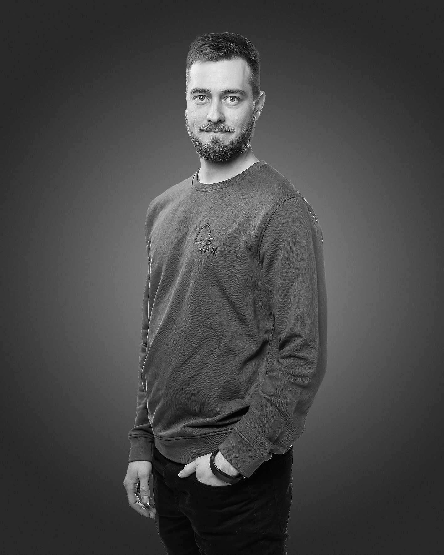 Parrakas henkilö seisoo yksinkertaista taustaa vasten yllään pitkähihainen teksturoitu neulepusero, käsi taskussa.