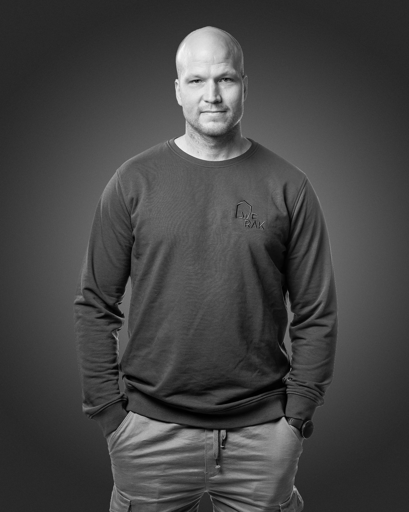 Henkilö, jolla on ajeltu pää ja yllään pitkähihainen collegepaita ja rennot housut, poseeraa tummaa taustaa vasten.