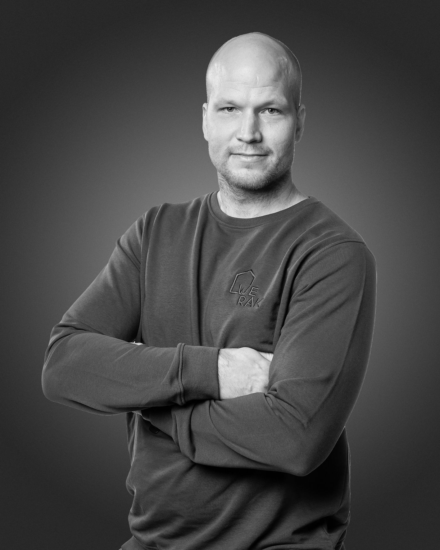 Henkilö, jolla on ajeltu pää ja yllään pyöreäkauluksinen neulepusero, seisoo kädet ristissä yksinkertaista harmaata taustaa vasten.