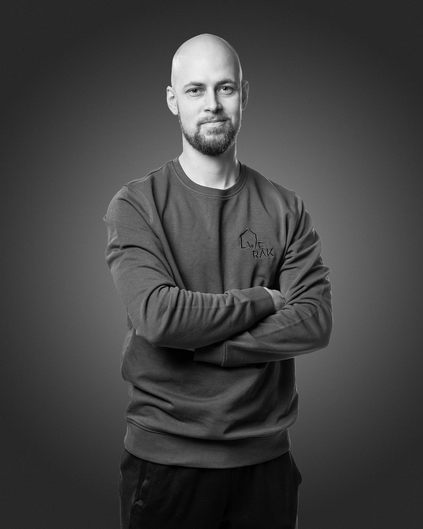 Hymyilevä, ajeltu pää ja parta, harmaa collegepaita yllään, mies seisoo kädet ristissä tummaa seinää vasten.