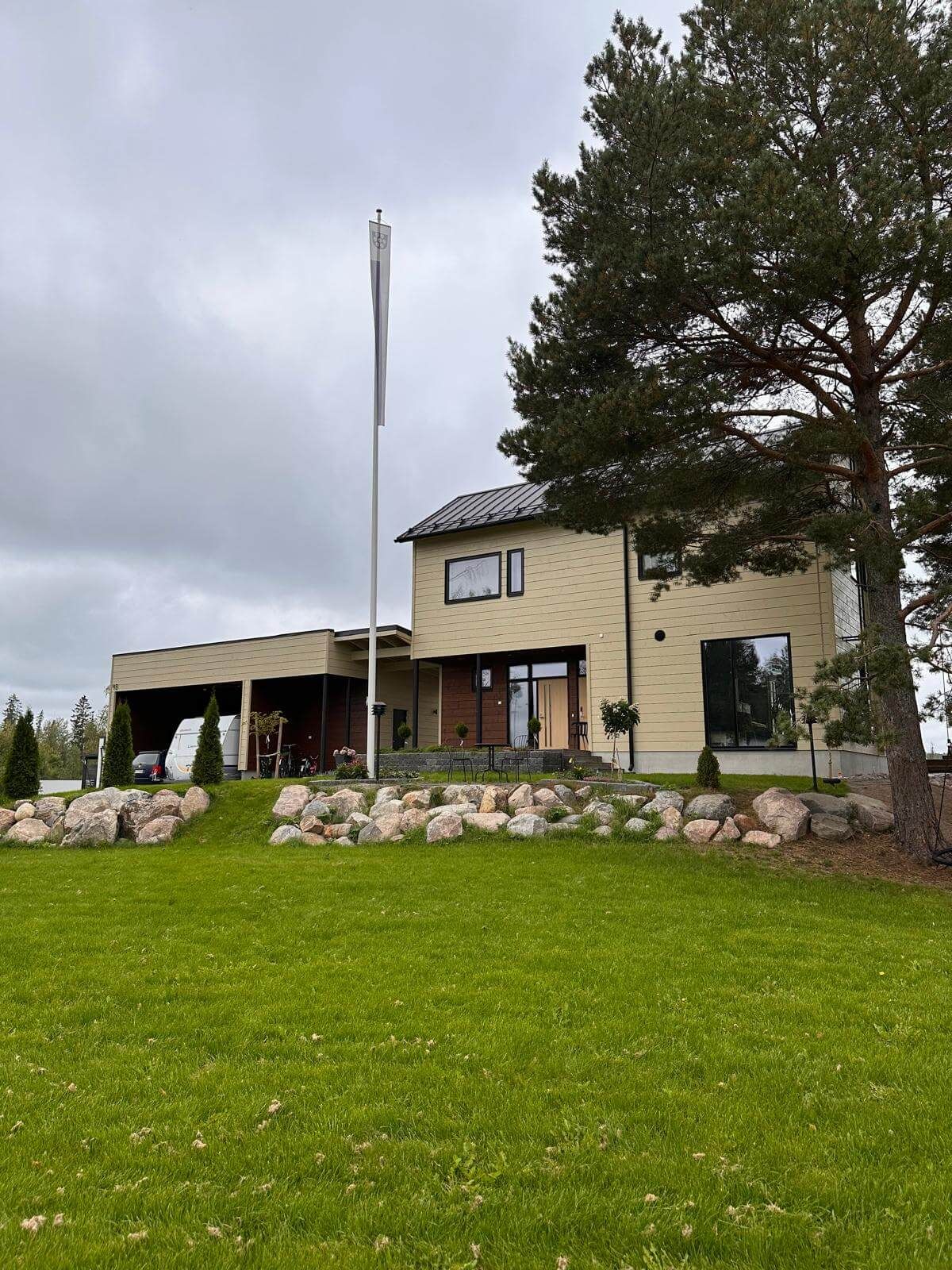 Vaalea kaksikerroksinen talo, jossa on autokatos, korkea lipputanko ja suuri mänty nurmikkoisella tontilla.